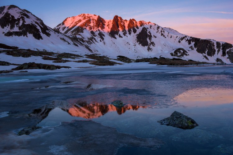 Suntem la inceputul primaverii sus in zona alpina din muntii Retezat, totul in jur este acoperit de un strat de zapada care s-a mai topit pe unele zone din crestele muntilor din jur si se vede roca de culoare maro inchis, doar varfurile muntilor sunt luminate de o lumina roz-portocaliu de rasarit, iar cerul este albastru senin, tot acest peisaj se oglindeste perfect intr-un lac din prim plan, doar in anumite portiuni se poate vedea, deoarece lacul este inghetat si doar in unele locuri nu are gheata pe suprafata acestuia, in coltul din dreapta jos un bolovan iasa din apa si seamana cao mica piramida.