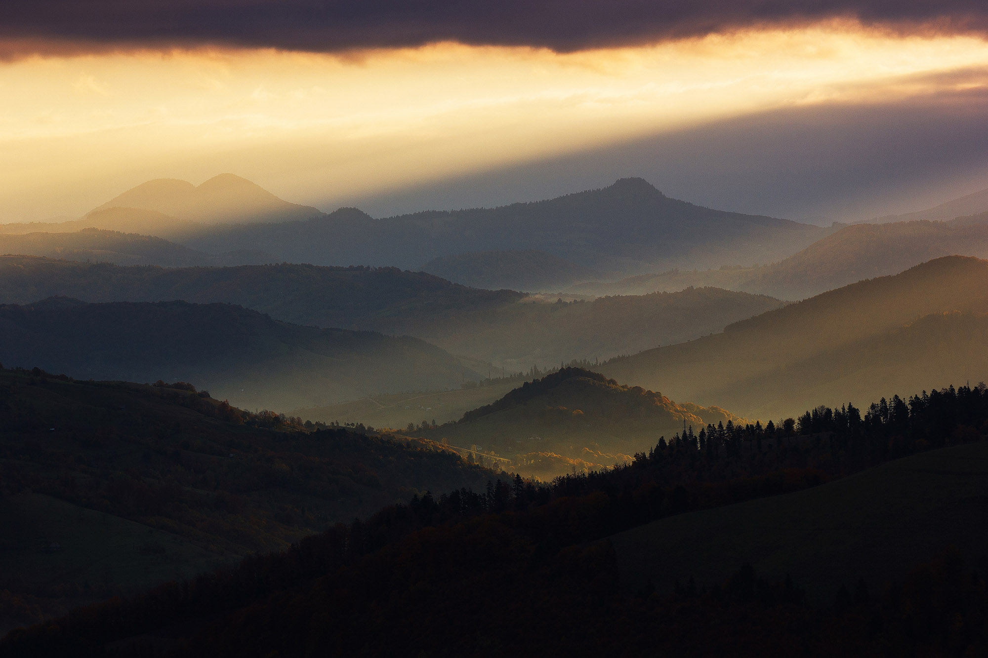 O imagine cu munti si dealuri cu o lumina magica de rasarit de soare cu raze care se revarsa peste peisajul din jur. In partea de sus avem niste nori negri iar dedesubt este cerul luminat si galben, lumina cade din lateral dreapta spre stanga jos.