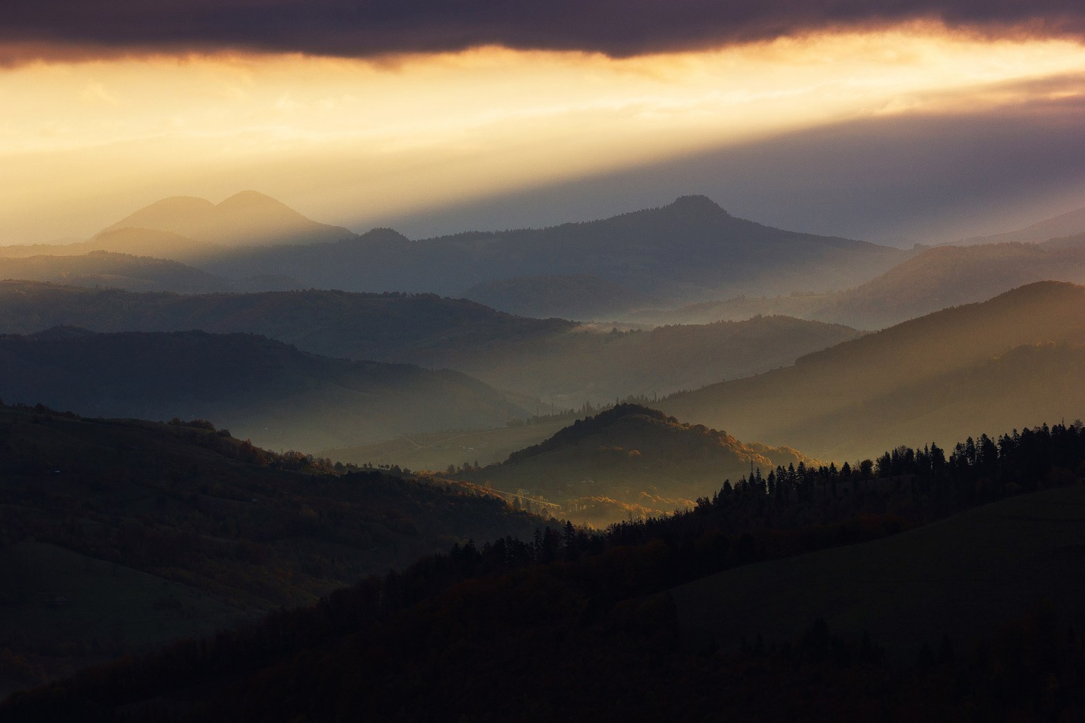 O imagine cu munti si dealuri cu o lumina magica de rasarit de soare cu raze care se revarsa peste peisajul din jur. In partea de sus avem niste nori negri iar dedesubt este cerul luminat si galben, lumina cade din lateral dreapta spre stanga jos.
