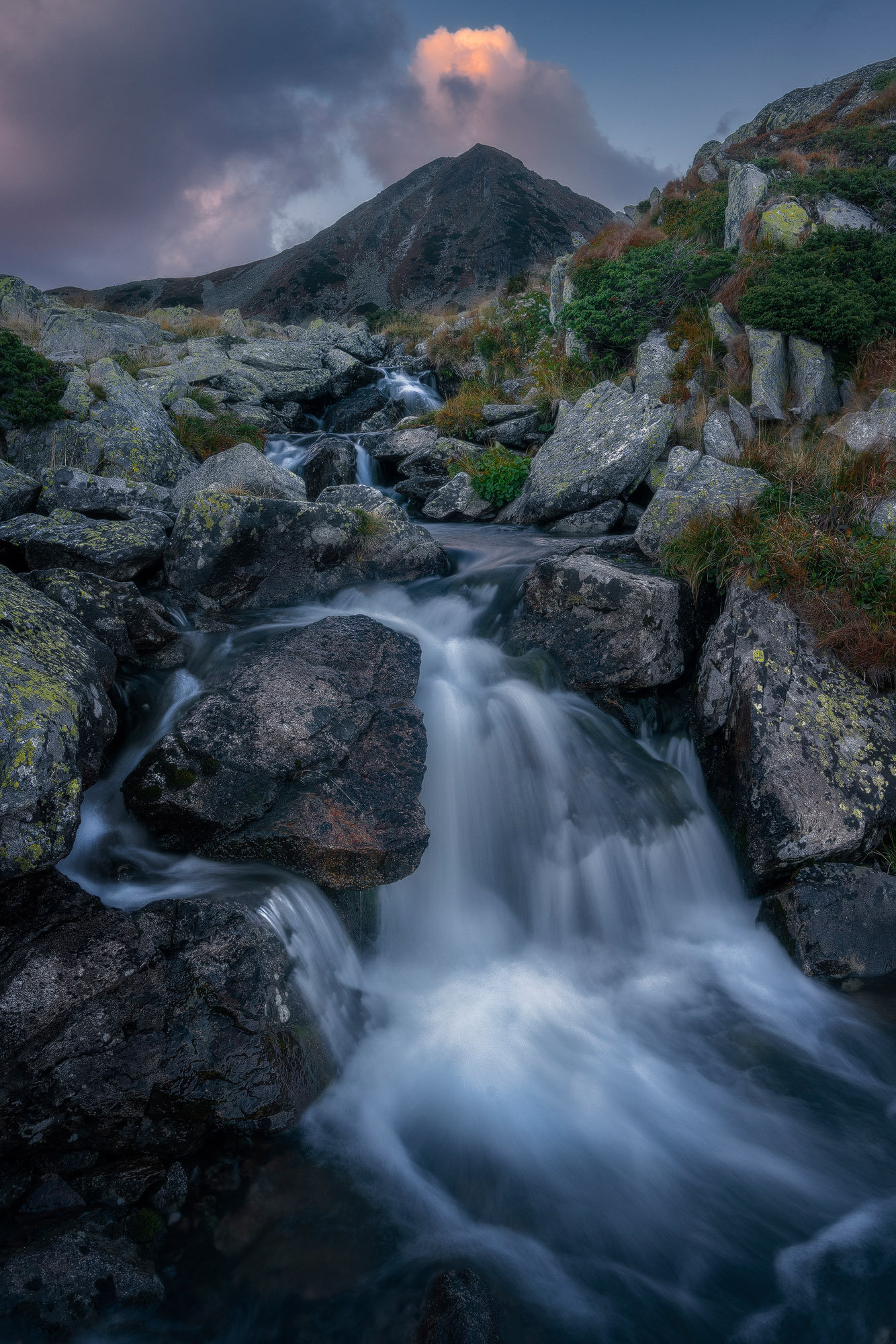 Parau de munte care curge in cascade printre bolovani, iar in fundal avem un varf de munte ascutit si impunator care are deasupra lui un nor aprins de ultima lumina a apusului.
