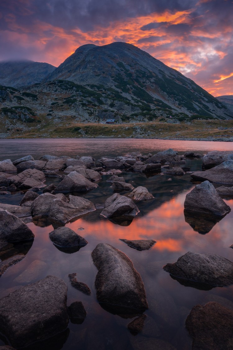 Un rasarit de toamna pe malului Lacului Bucura, avem un cer acoperit cu nori colorati de lumina rasaritului in nuante de rosu aprins, portocaliu, roz si mov deasupra unui varf de munte, iar toata aceasta lumina este reflectata pe suprafata lacului printre pietrele prezente la marginea acestuia. Undeva in centrul imaginii chiar pe directia varfului de munte , putin deasupra lacului avem o mica casuta, de fapt este un refugiu montan, se numeste refugiul Bucura.