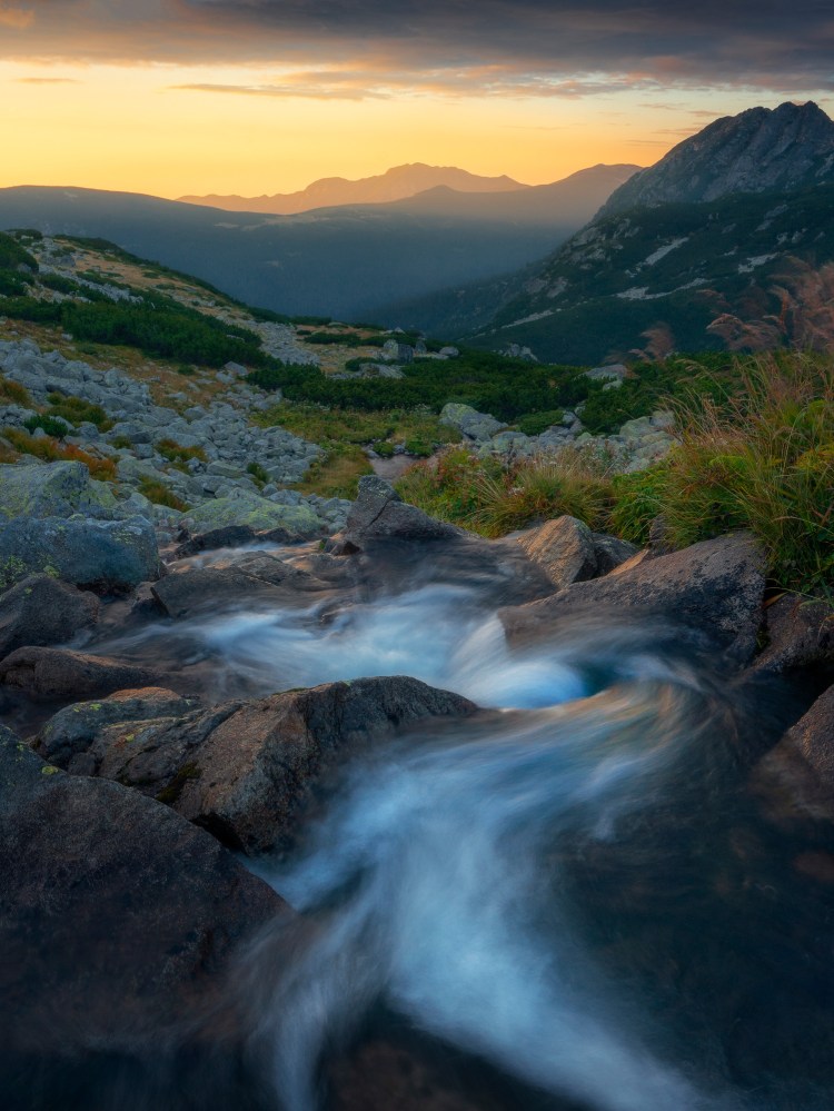 Rau de munte serpuind printre bolovani colorat cu o tenta albastruie-aurie si care ne poarta privirea spre muntii din fundal si spre cerul auriu colorat de lumina rasaritului. Un rasarit de toamna spectaculos.