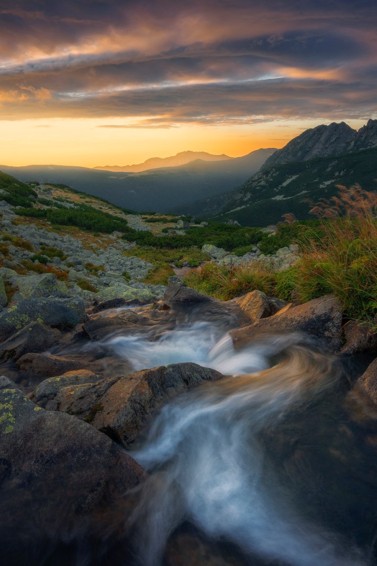 Rasarit in munti, Avem in fata un rau care curge serpuind printre bolovani si ne poarta privirea spre munti si spre un cer cu nori de o culoare deosebita.