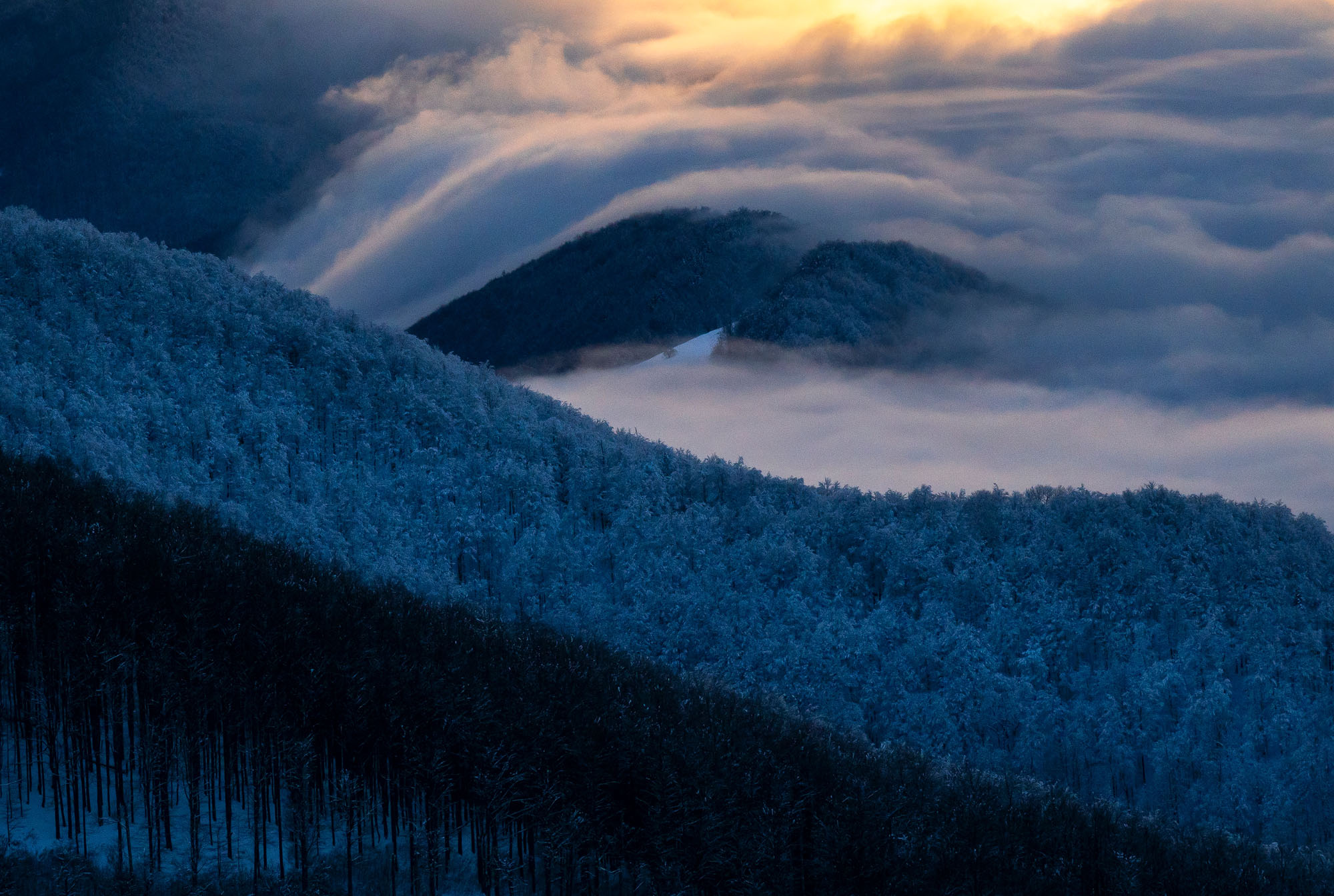 A serene landscape captures snow-covered hills beneath a blanket of rolling clouds, with the setting sun casting a warm glow in the sky. The contrast between the cold blue tones of the snow and the golden hues of the sunset creates a mesmerizing scene.