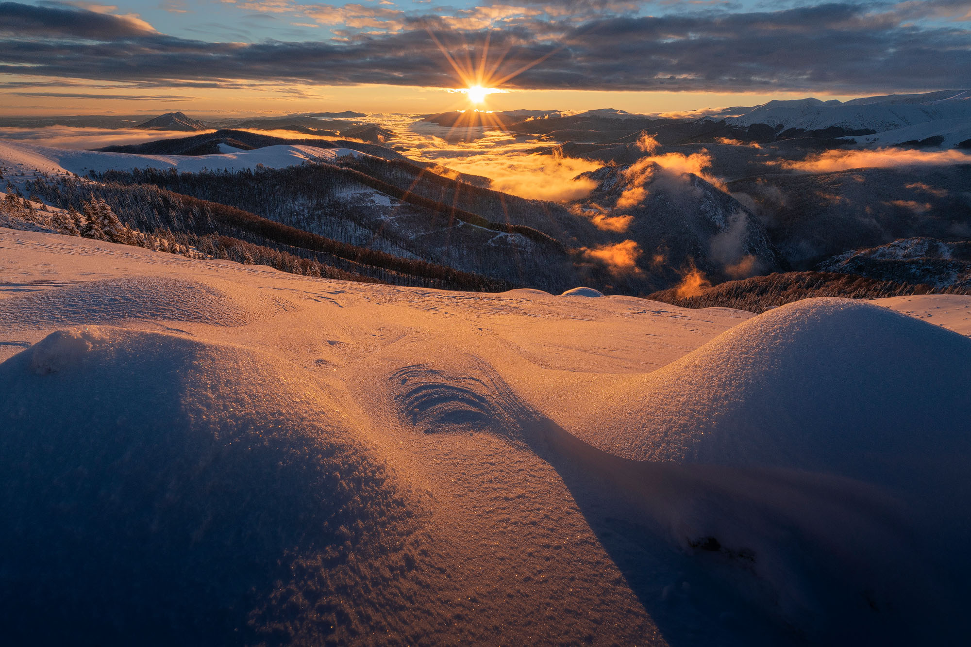 Golden sunlight bathes the snow-covered mountains as the sun sets behind a distant horizon. Soft clouds linger in the valleys, creating a serene contrast between the glowing sky and the white landscape. Image made in Valcan Mountains, Romania.