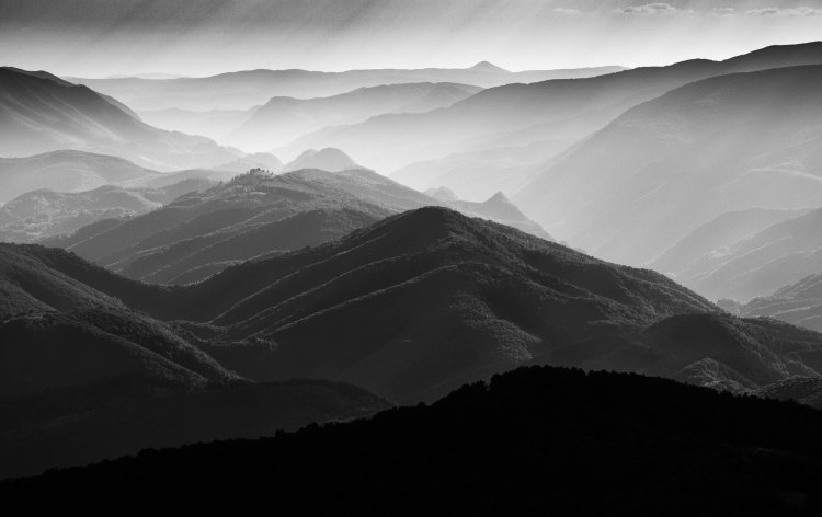 Layers of rolling mountains stretch toward the horizon in soft grayscale, fading into a dreamy haze that blurs the boundary between earth and sky. Each ridge is gently defined by shifting light, creating a mesmerizing rhythm of shadow and highlight across their forested slopes. The distant peaks dissolve into mist, giving the scene an ethereal sense of depth and quietude. Above, a pale sky glows softly, its light filtering down like a whisper over the vast landscape. The overall mood is serene and contemplative, as though time itself has slowed to honor the stillness of the mountains.